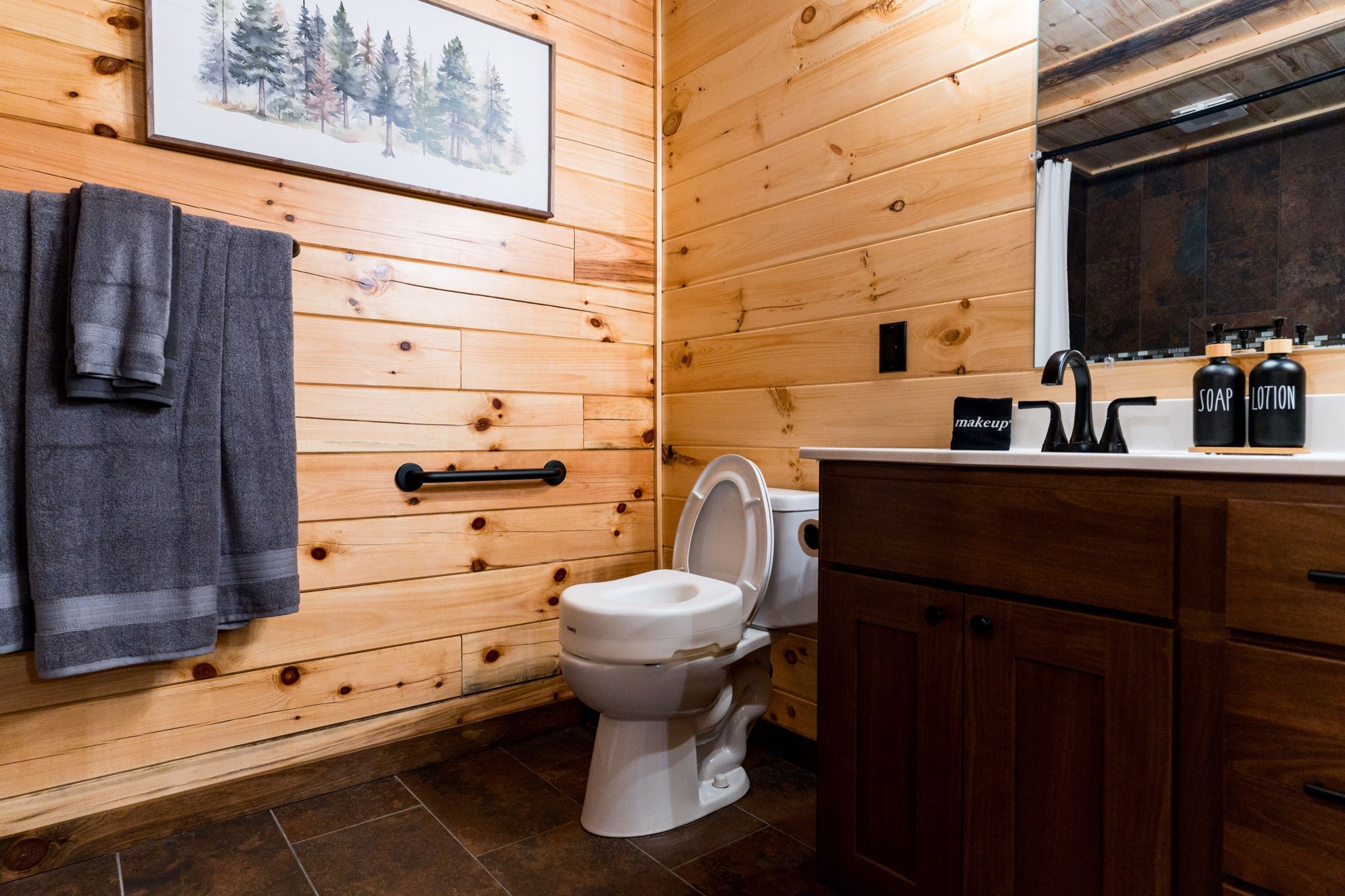 The toilet in the accessible bathroom with a raised seat fitted and a black grab bar on the pine wall beside it.