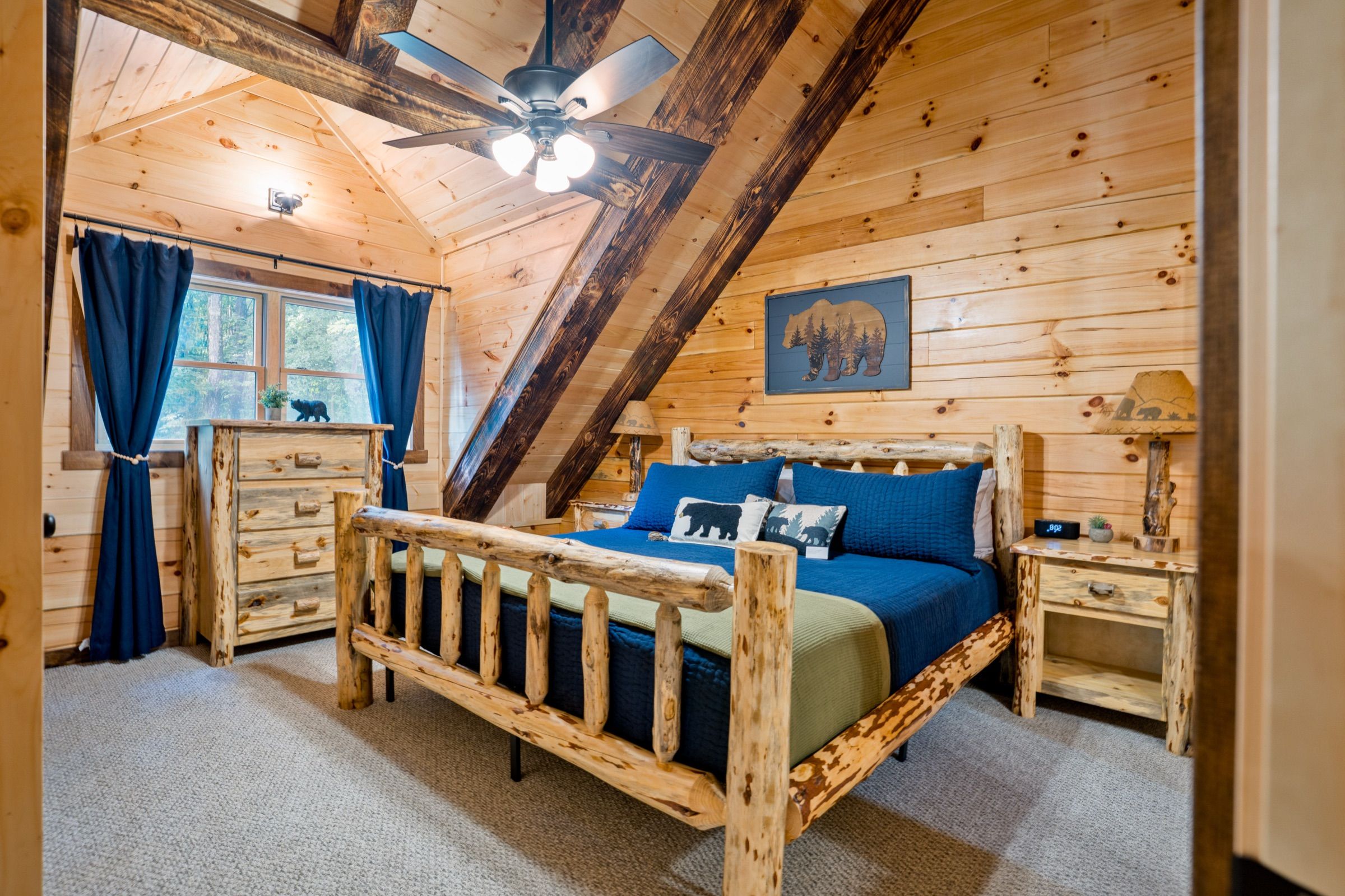 King bedroom with a log-frame bed, blue quilt, and charred timber beams meeting in a vaulted pine ceiling; a window with navy curtains looks into the trees.