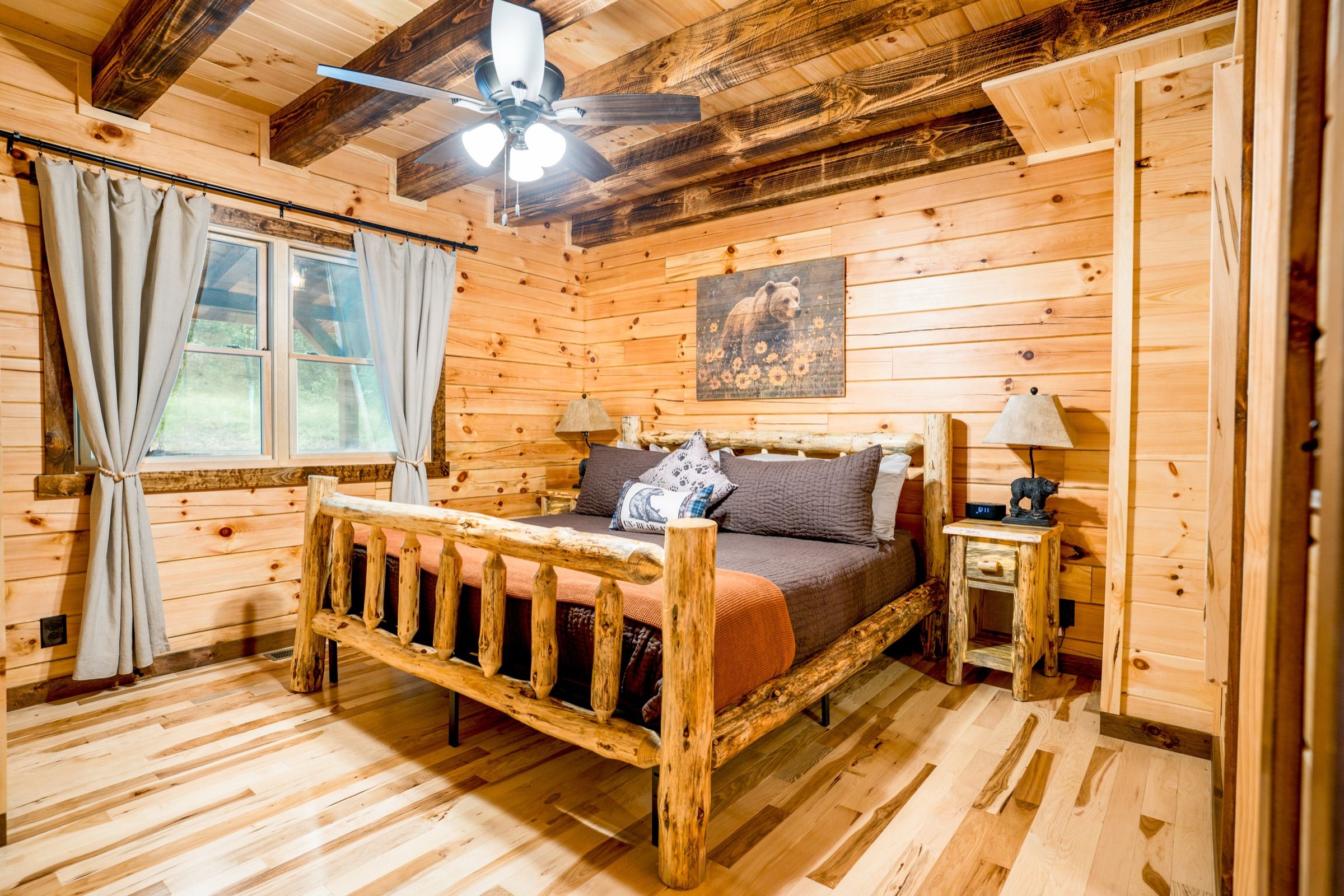 The main-floor king suite: a log-frame king bed dressed in brown and rust, pine walls, dark beams, and a bear print above the headboard. Its door and bathroom door are 36 inches wide.