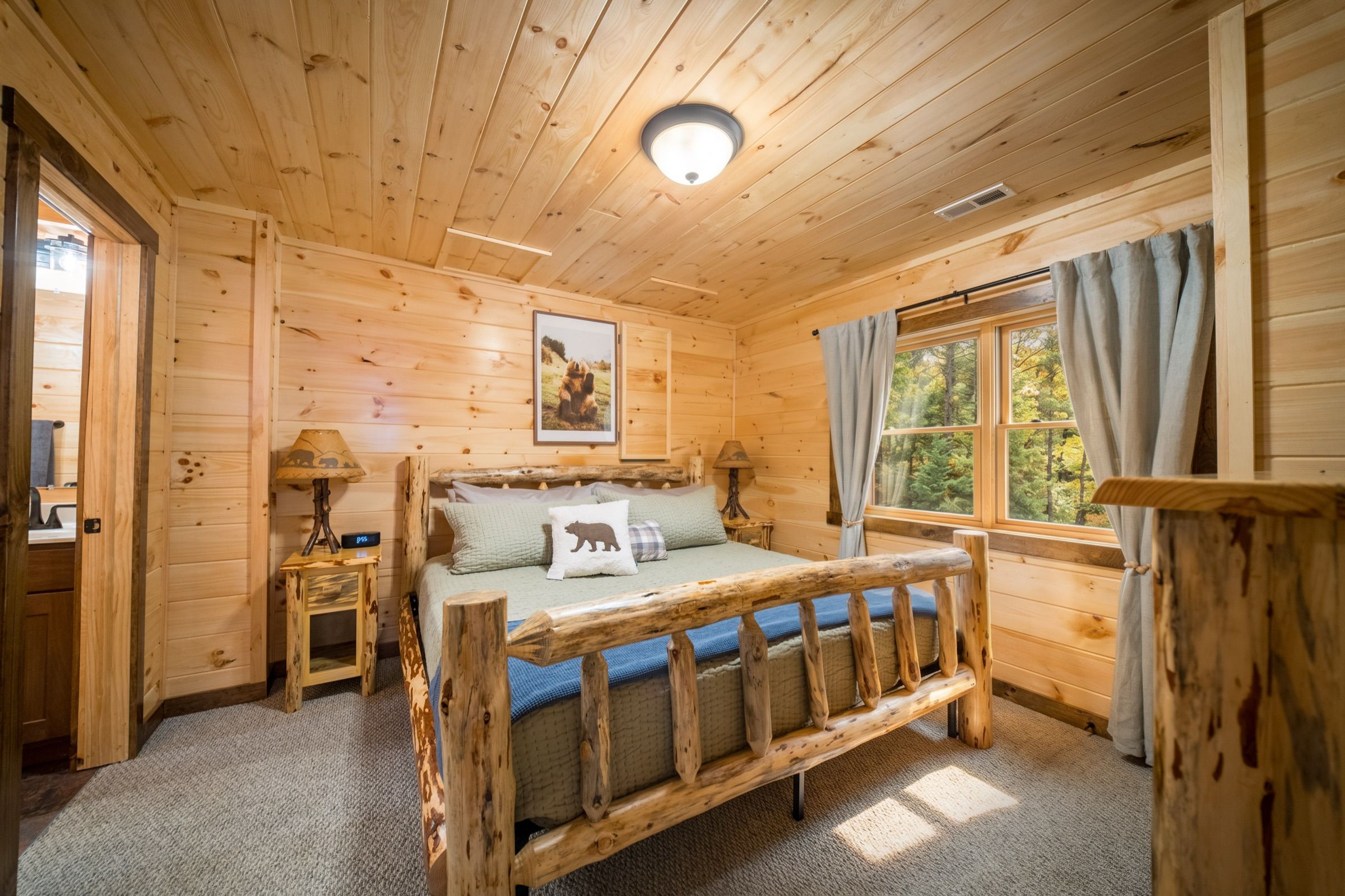 King bedroom with a log-frame bed in sage and blue, pine walls, and a window full of afternoon trees; the door to its bathroom stands open.