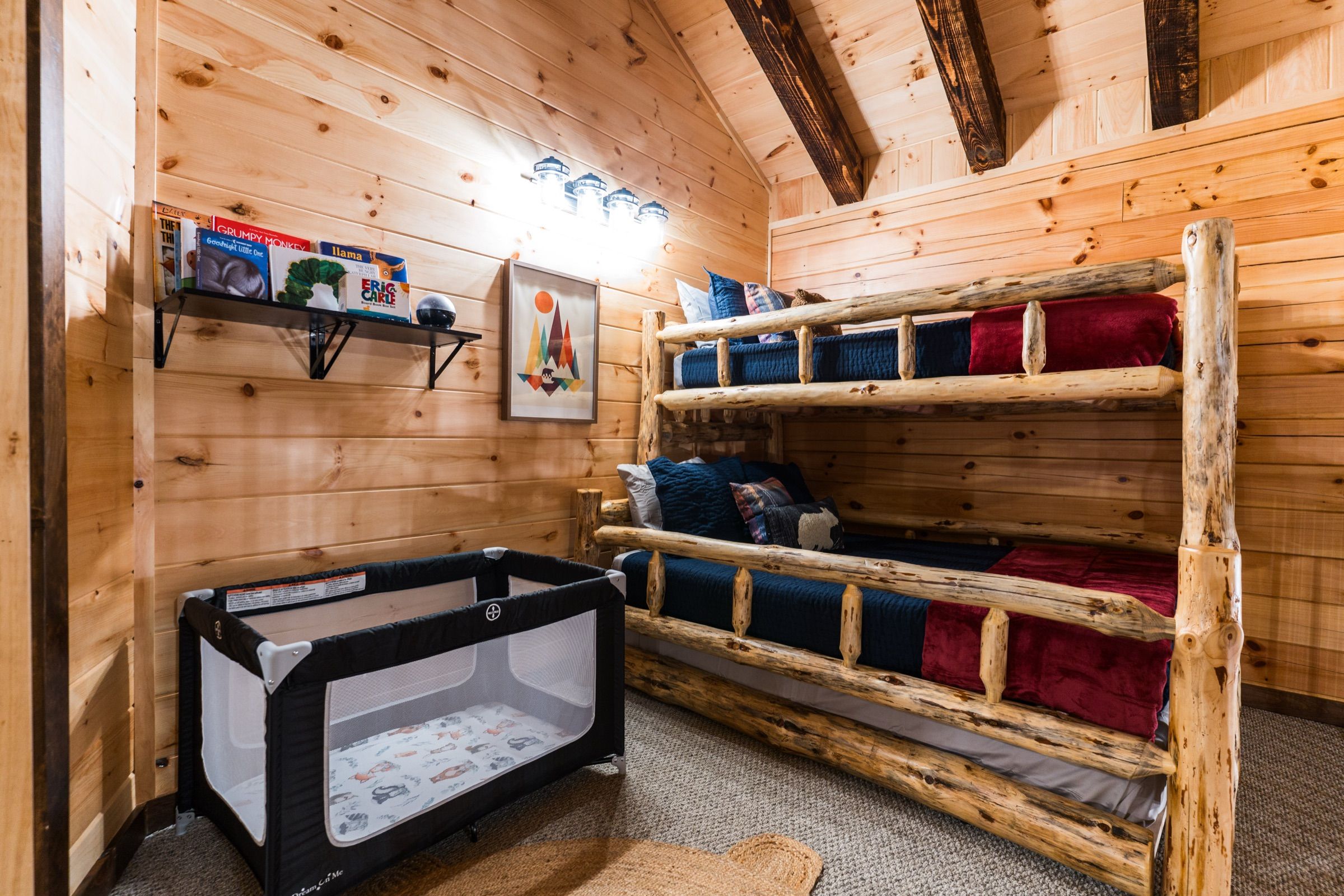 The bunk loft: log-frame bunk beds with navy quilts and red blankets, a shelf of picture books, and a Pack 'n Play set up in the corner.