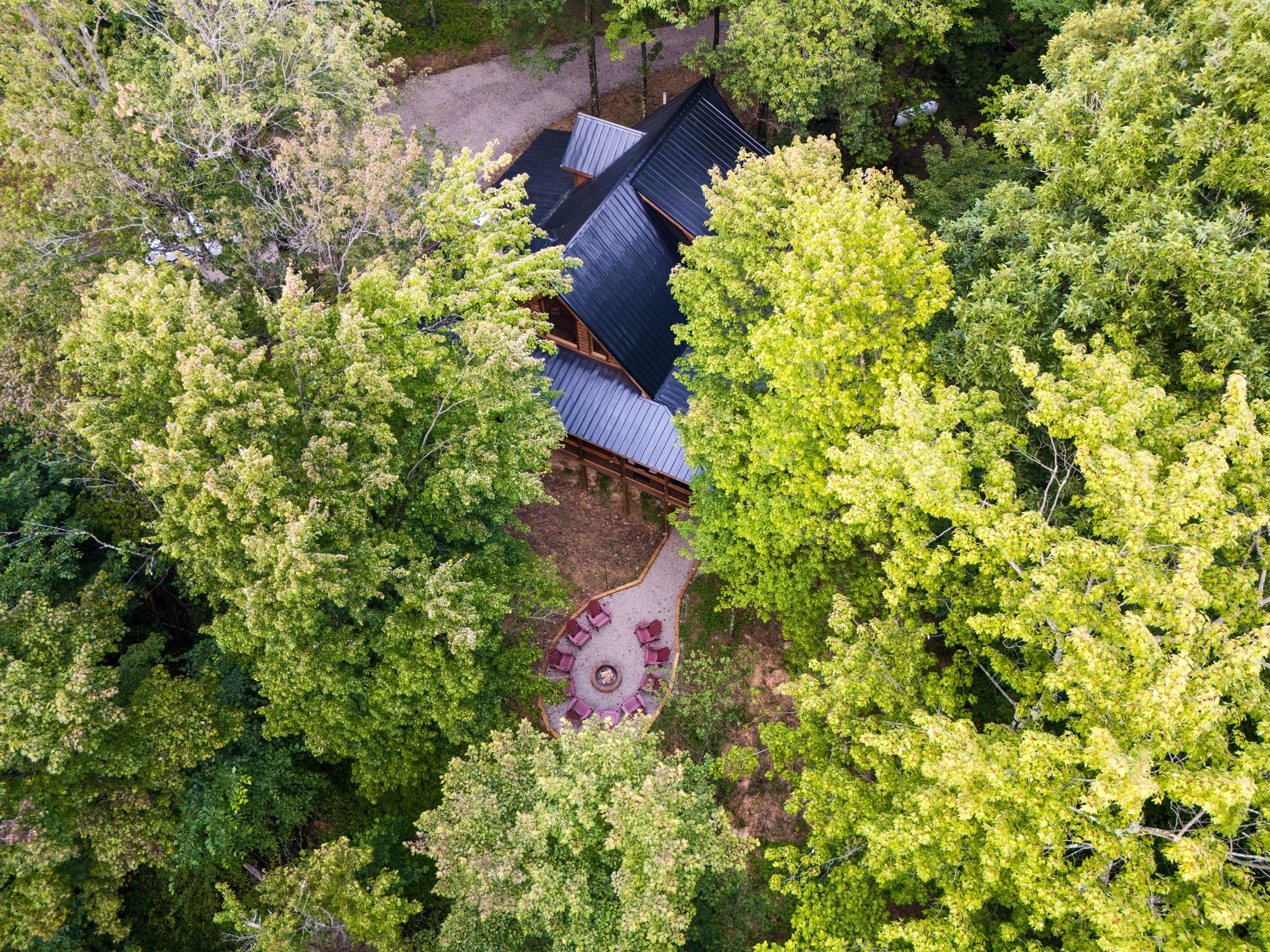 Drone view straight down onto Sleepy Bear Lodge: a black metal roof in dense green tree canopy, with a gravel path leading to a ring of red Adirondack chairs around a stone fire pit.