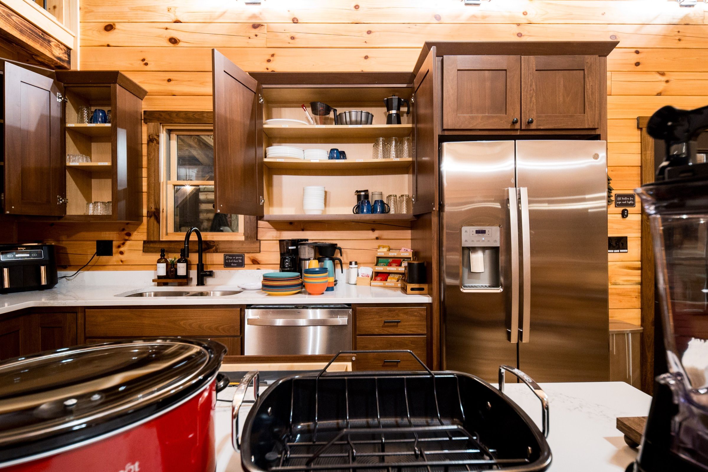 Kitchen with walnut cabinets, white quartz counters, a stainless French-door refrigerator, two coffee makers, and open shelves of stacked dishes.