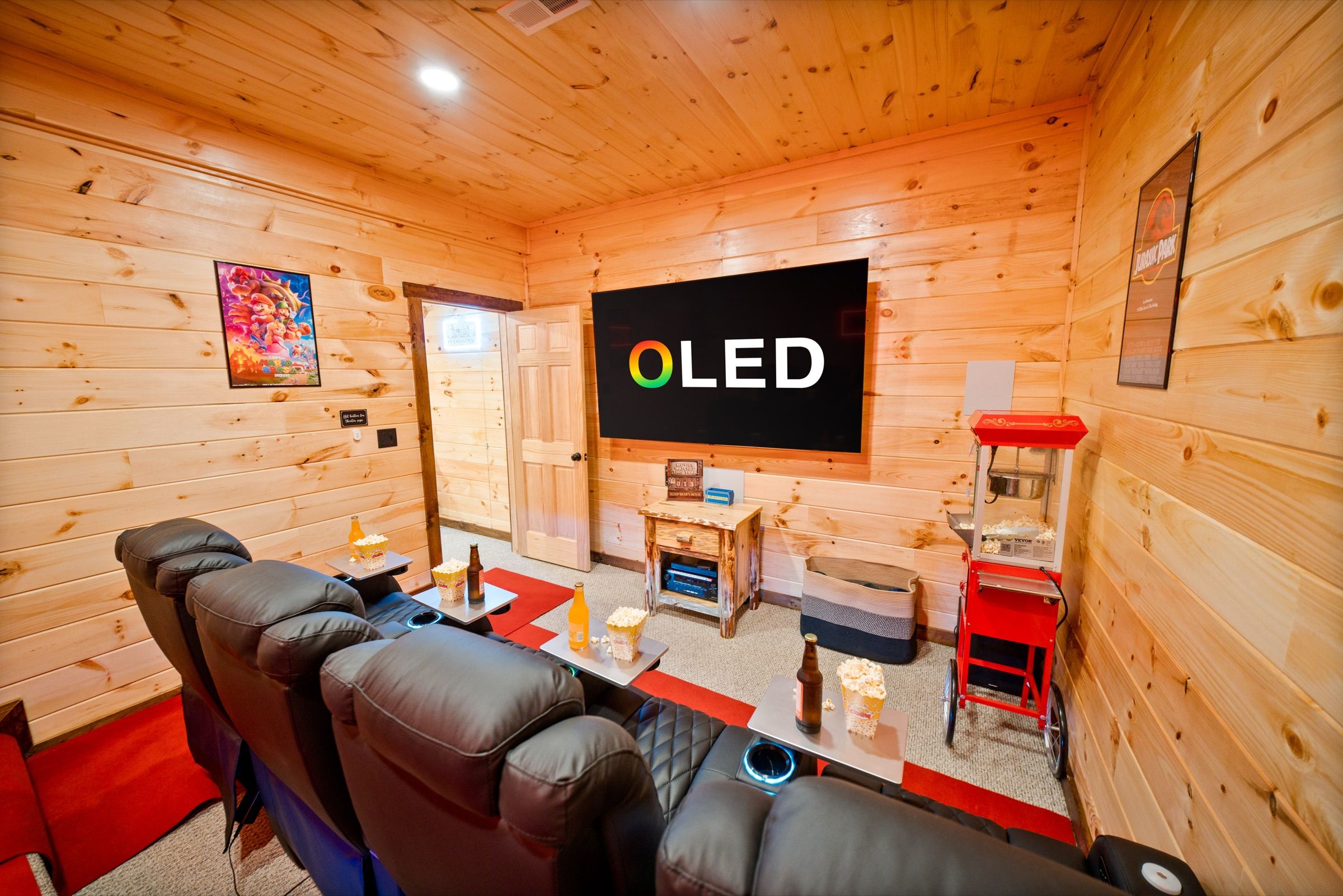 Home theater with a row of black leather recliners, a large OLED screen on a pine wall, a red popcorn machine, and movie posters.