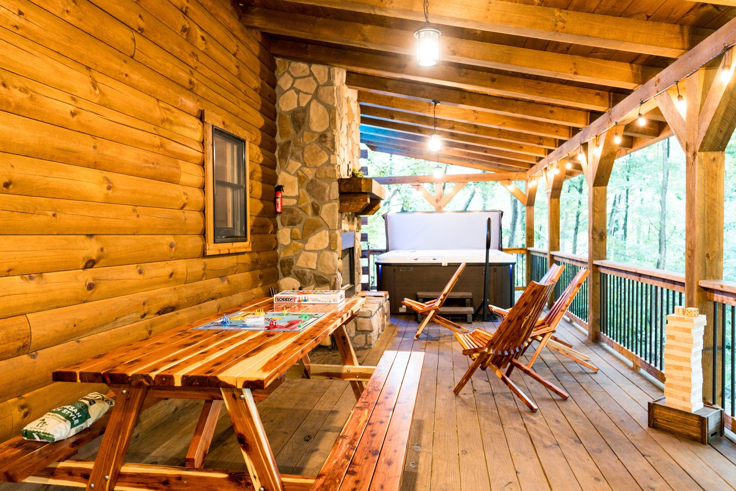 The covered deck: a cedar picnic table set with board games, folding chairs, an outdoor stone fireplace, the hot tub under its cover, and string lights along the rail.